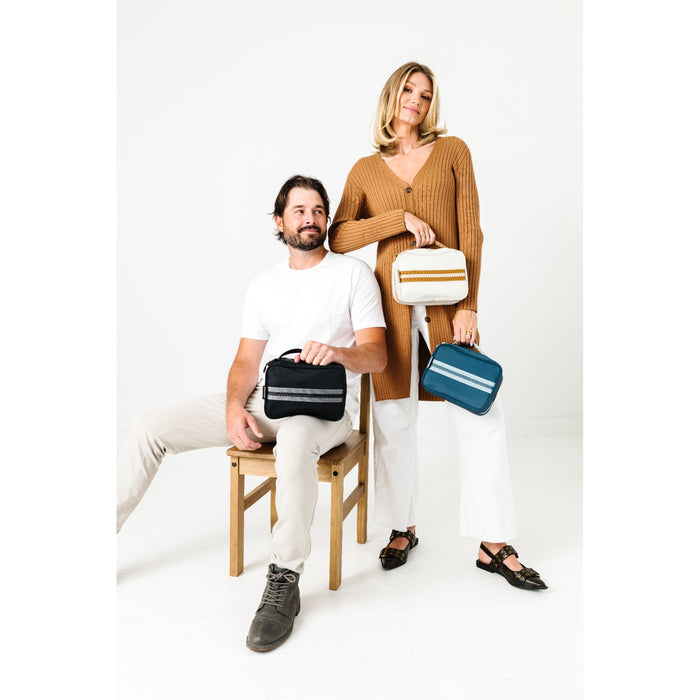 A man and a woman pose with three small rectangular handbags in a bright studio setting. The man sits on a wooden chair holding a black bag with white stripes, while the woman stands behind him holding a cream and gold bag and a blue bag with white stripes.