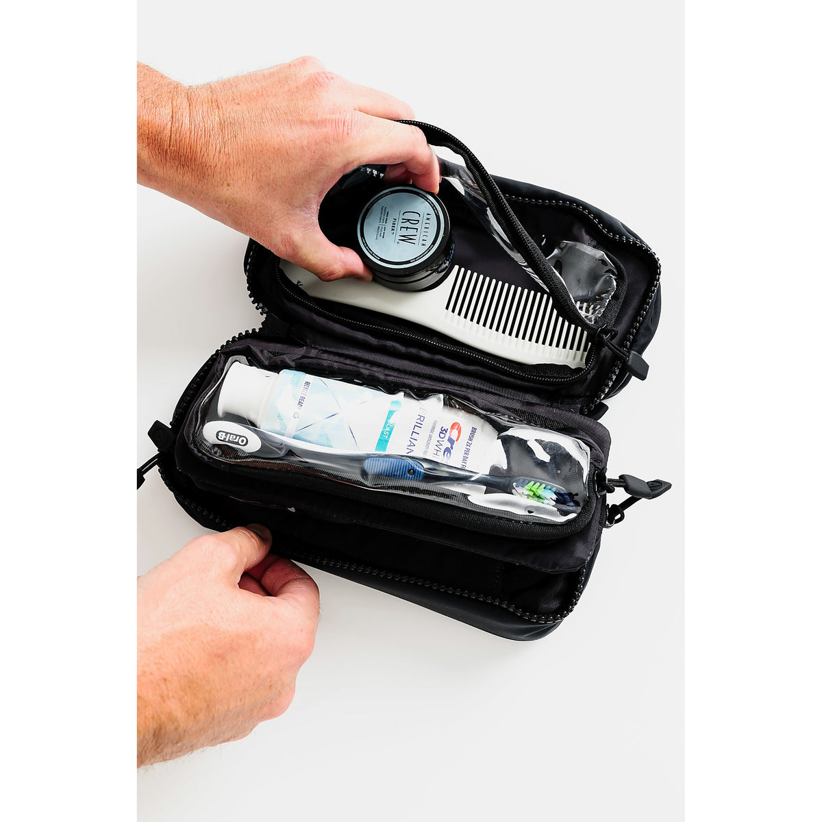 A black zippered travel organizer case shown open on a white background, with a person's hands placing and removing toiletry items including a round container, comb, toothbrush, and toothpaste tube.