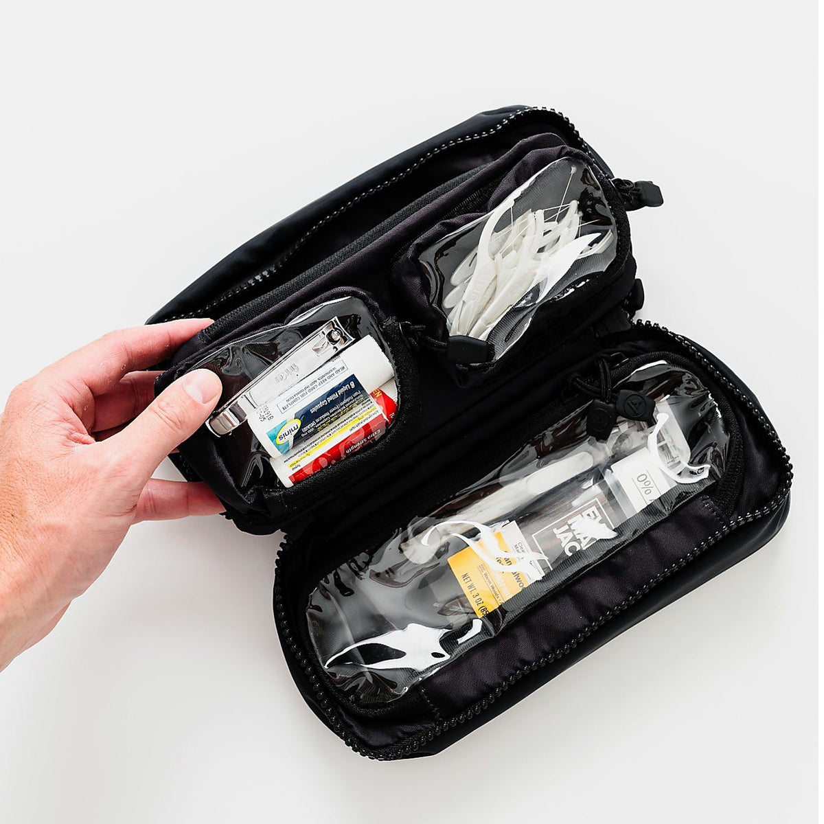 A black zippered medical or first-aid kit pouch shown open on a white background, with a hand holding one side to display the interior compartments containing packaged supplies.