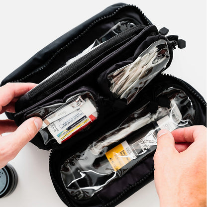 An open black zippered medical or first-aid pouch with clear plastic compartments containing various small medical supplies, shown with two hands reaching in on a white background.