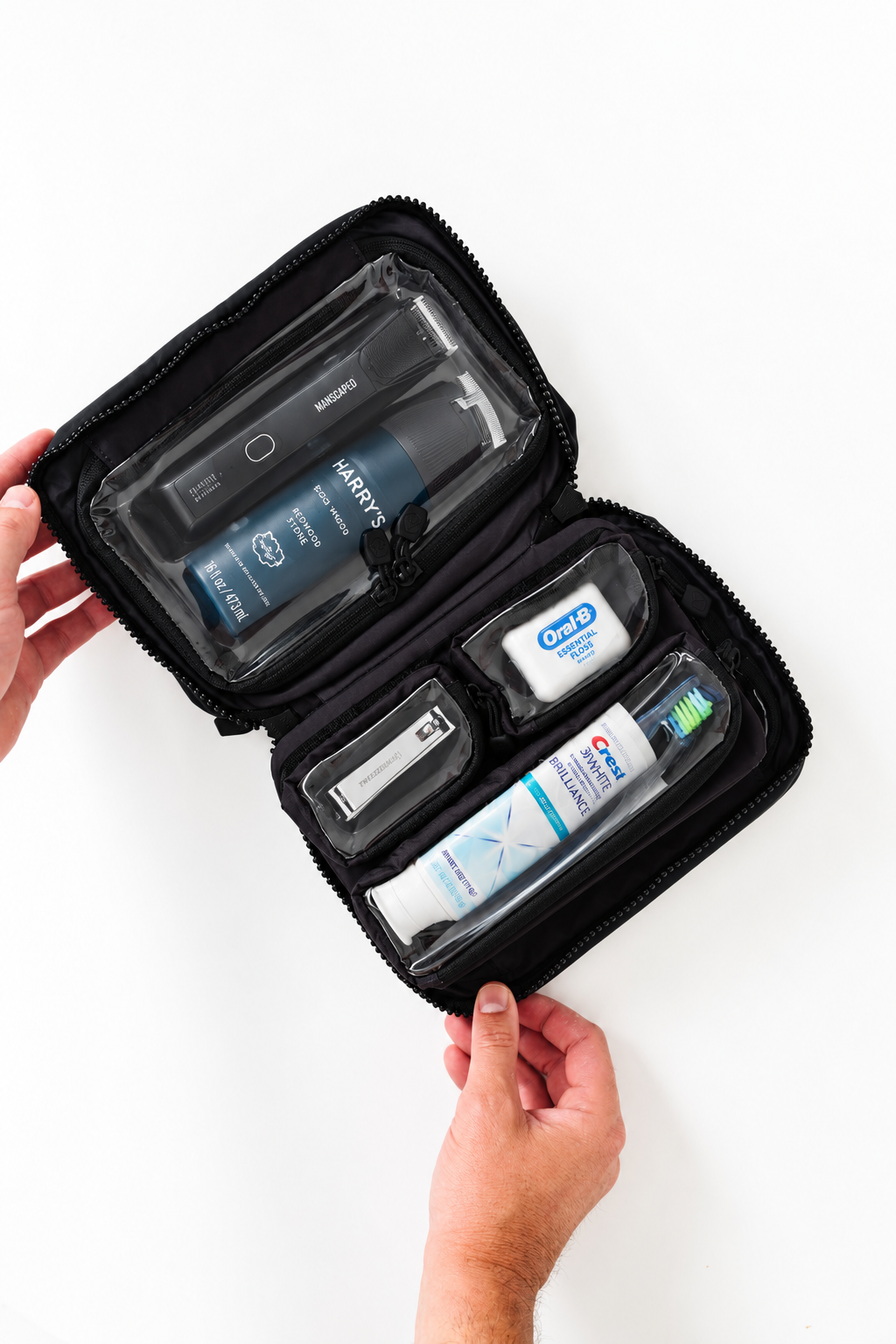 A black zippered travel toiletry organizer case held open by two hands, showing several toiletry items inside including a shower head, toothbrush, toothpaste, and other small grooming products, on a clean white background.