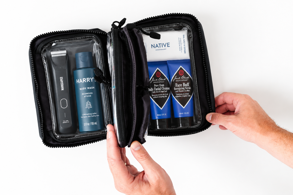 A black zippered travel toiletry case opened flat on a white background, containing a mirror, a blue body wash bottle labeled HARRY ROYCE, and two blue face care tubes labeled NATIVE and The Ordinary. Two hands are holding the case open.