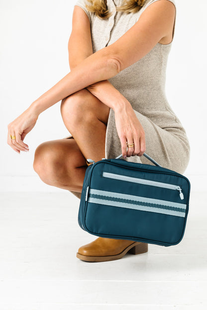A woman in a beige sleeveless dress crouches while holding a teal toiletry bag with two white horizontal stripes and a top handle, against a clean white studio background.