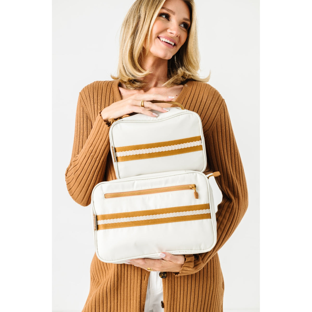 A woman in a brown ribbed cardigan holds two white diaper pail bags with mustard yellow and white stripes against a plain white background.
