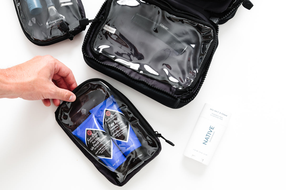 Three black zippered toiletry bags with clear plastic windows are arranged on a white background. A hand is holding one bag open, and a white tube labeled NATIVE is placed beside it.