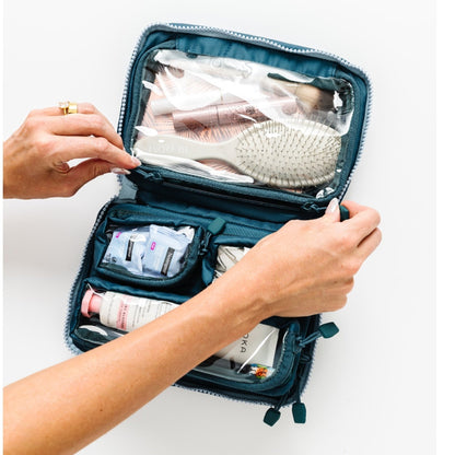 An open teal blue travel toiletry case with clear zippered compartments and internal pockets, shown being organized with hand and arms against a plain white background.
