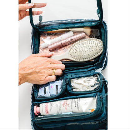 An open teal makeup and travel organizer bag with clear zippered compartments, shown with hands placing and retrieving items such as a hairbrush, lip gloss, and skincare tubes. The shot is cropped to focus on the bag and the hands interacting with it against a plain white background.