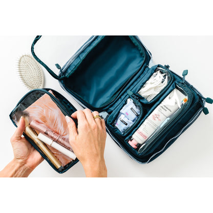 An open dark teal makeup and travel organizer bag shown from above, with hands holding a smaller matching pouch containing cosmetics. The main bag has clear zip compartments containing cotton pads, a tube, and other travel items on a white background.