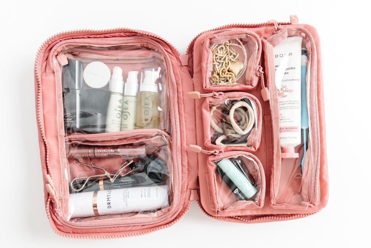 An open pink zippered travel organizer bag with clear pockets containing various toiletries and accessories, photographed from above on a white background.