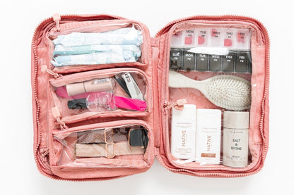 An open pink makeup bag filled with cosmetics and travel-sized items, photographed from above on a clean white background.