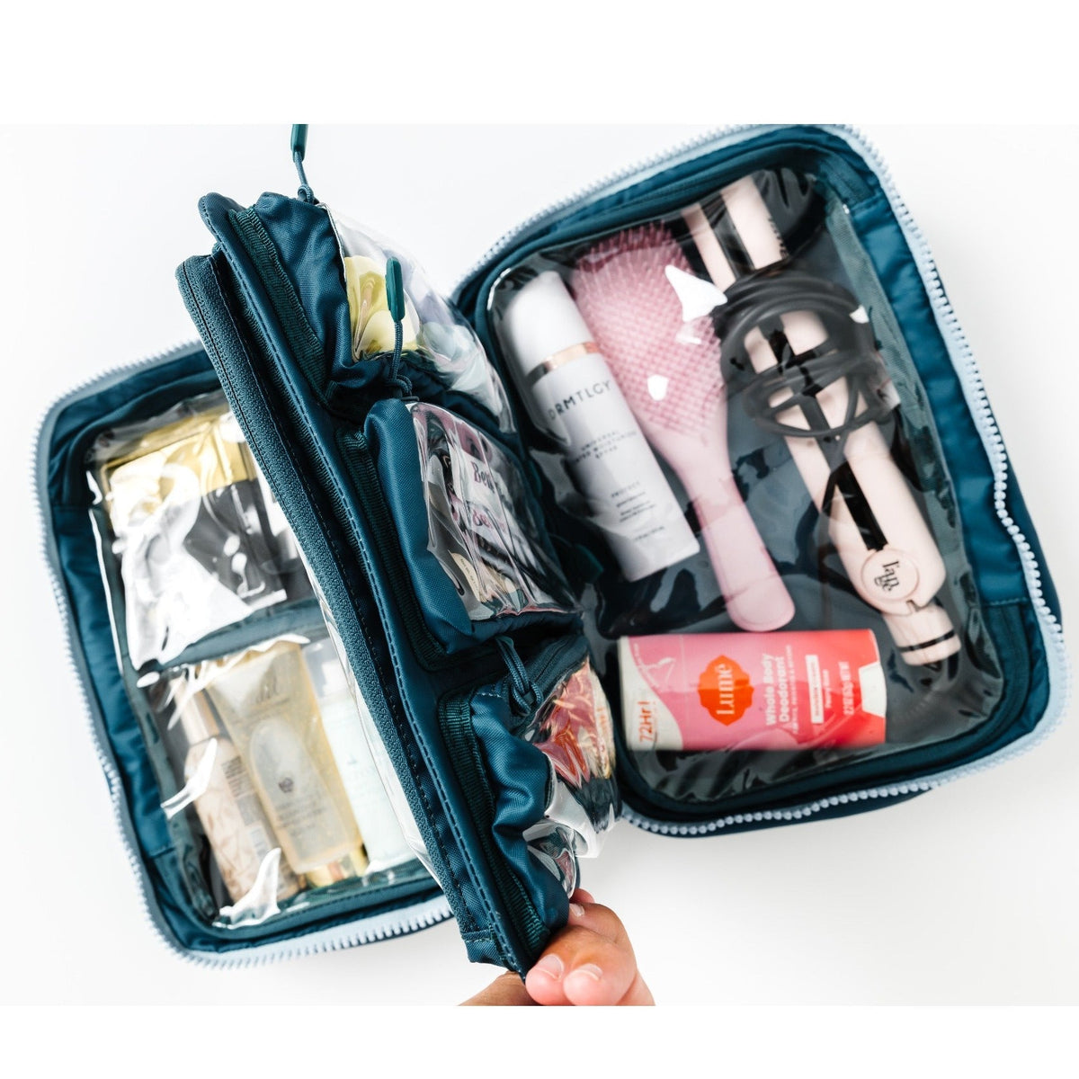 An open dark teal zippered travel organizer bag with clear pockets, shown on a white background with a hand holding the top edge. The bag contains various toiletries and travel items.