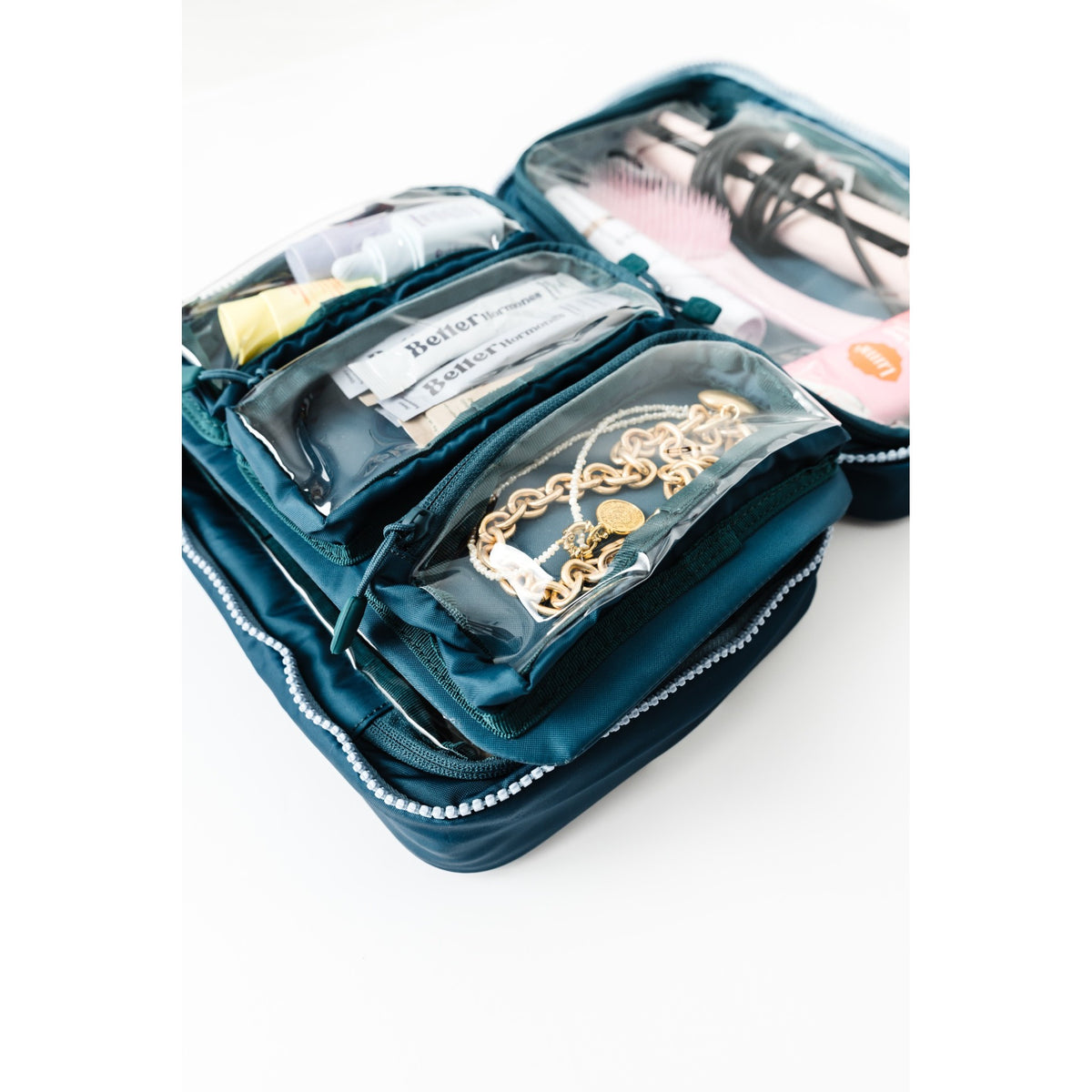 An open dark teal nylon travel organizer pouch with clear zippered pockets, shown on a white background. The pouch contains various toiletries and accessories, including a gold chain necklace, a white packet labeled Better Days, and other small items.