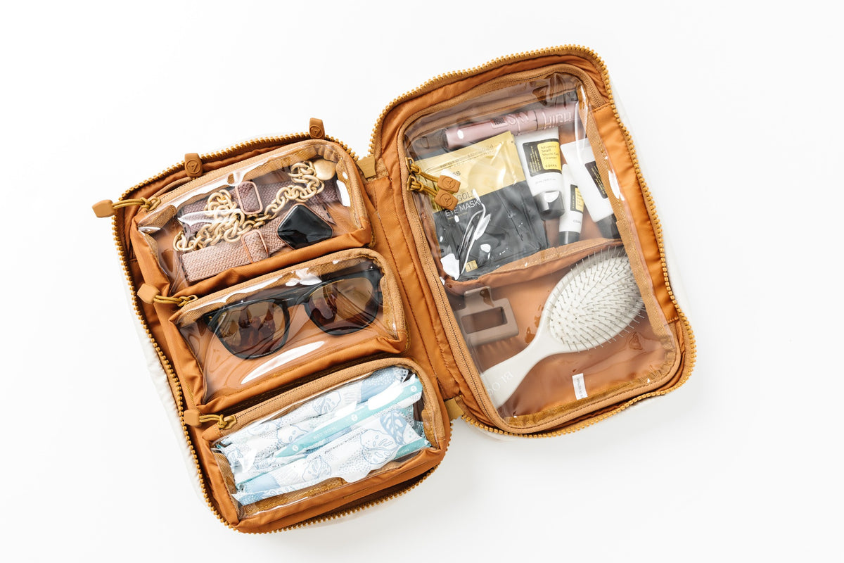 An open tan zippered travel organizer bag with clear plastic compartments, shown from above on a white background. The bag is filled with various travel items including sunglasses, a hairbrush, a gold chain, and a face mask.
