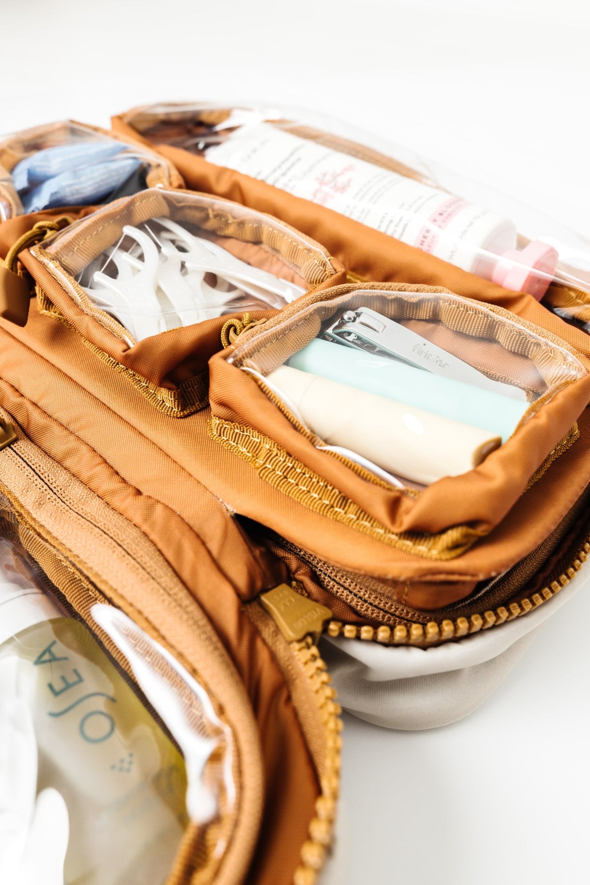 An open tan canvas toiletry bag with multiple clear zippered pockets containing personal care items, photographed on a white background.