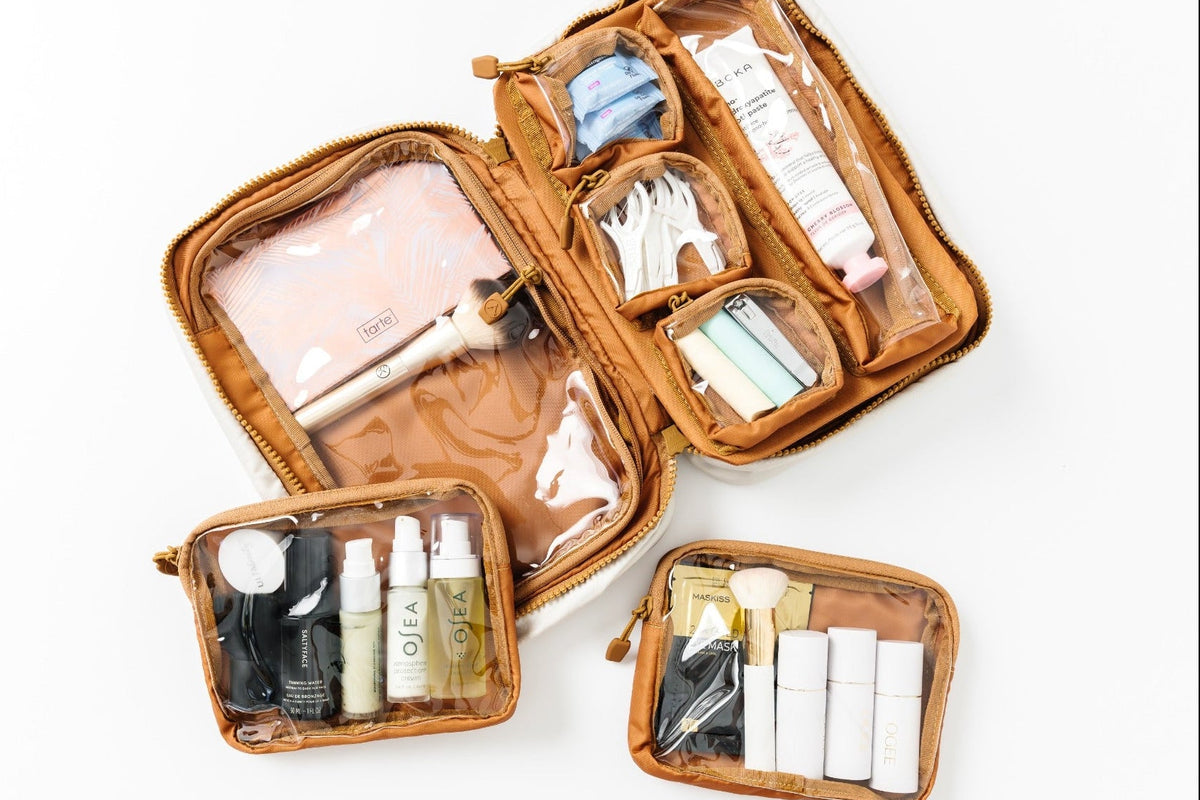 Three tan zippered travel makeup bags arranged on a white background, with the main bag open to show internal compartments and two smaller bags beside it, each containing assorted toiletries and cosmetics.