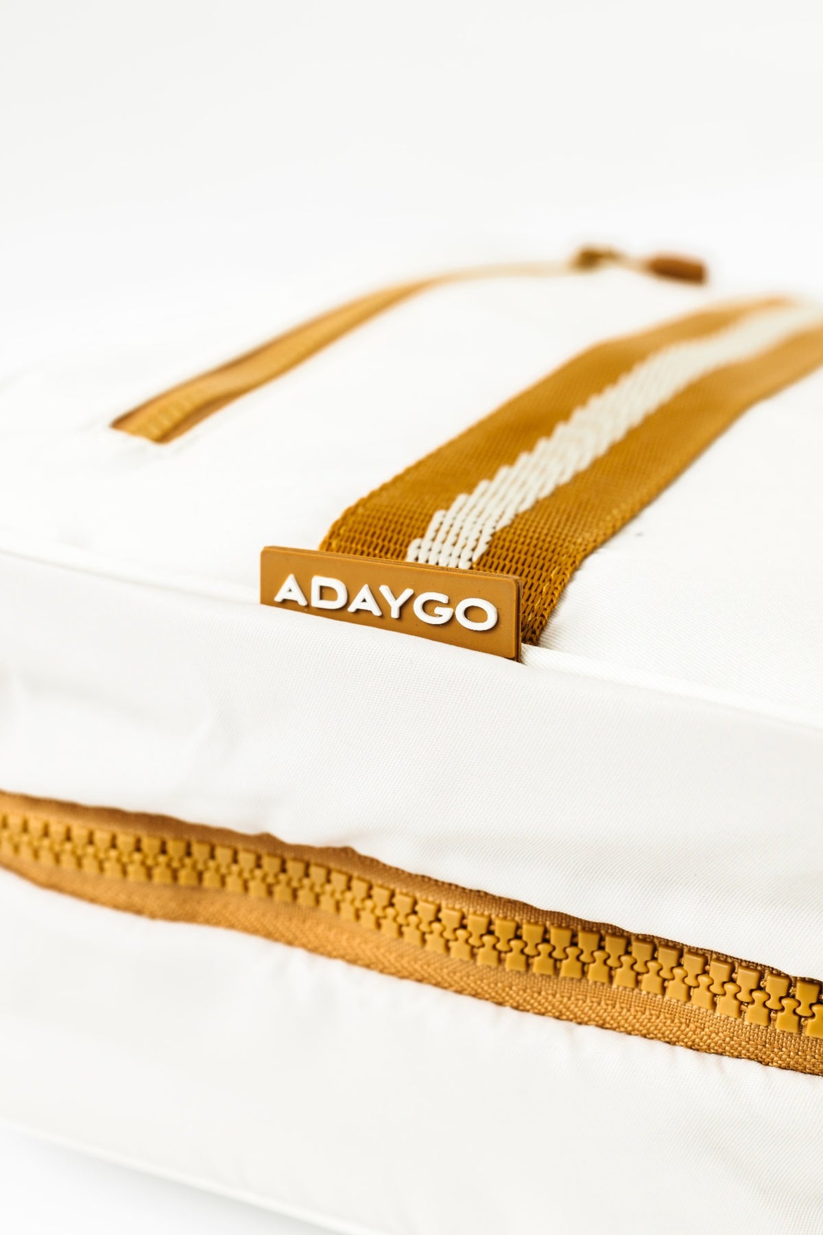 A close-up of a white soft-sided bag with a mustard yellow zipper, a matching yellow webbing strap, and a small brown brand tab reading ADAYGO.
