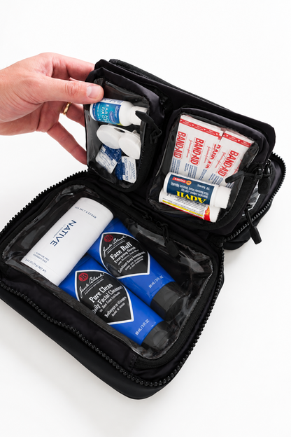 An open black zippered travel first-aid kit pouch on a white background, showing organized compartments with bandages, gauze, and skincare items including a NativE tube and blue Pure Encapsulations tubes. A hand is holding the top edge of the pouch.